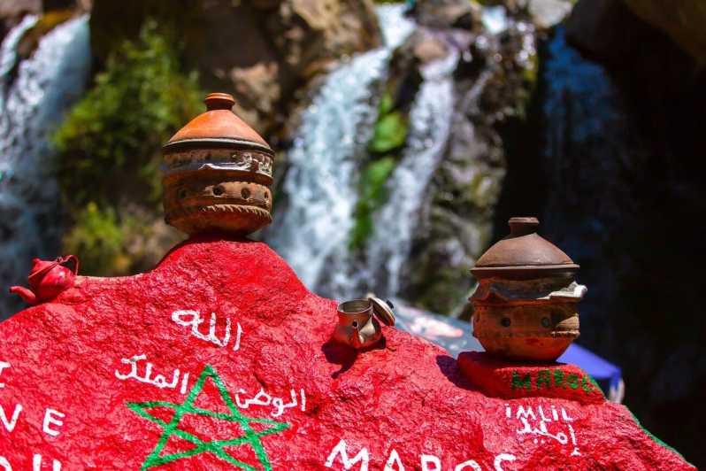 Close-up of Moroccan cultural symbols, including traditional tagine pots and a painted rock with Arabic inscriptions, set against the backdrop of a cascading waterfall in the Atlas Mountains during the Marrakech: Atlas Mountains, Berber Villages & Waterfall Tour.